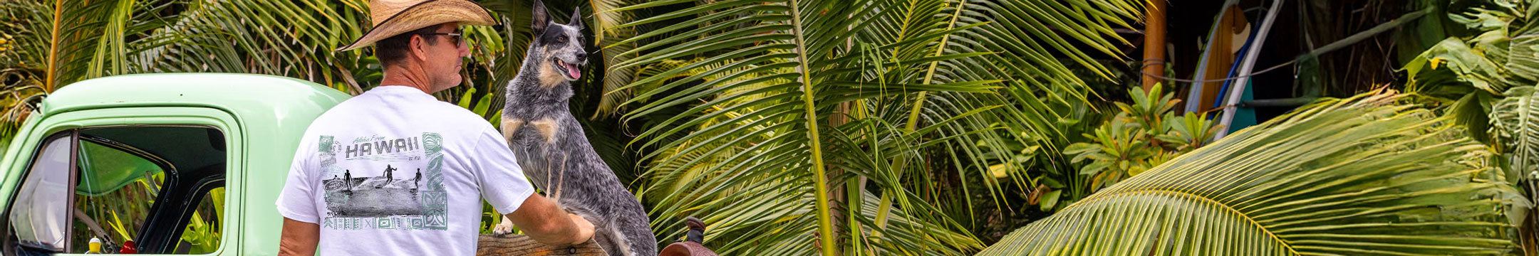 Man with a dog standing next to a vintage truck in a tropical setting Wearing a Crazy shirt with a surfing design on it. 