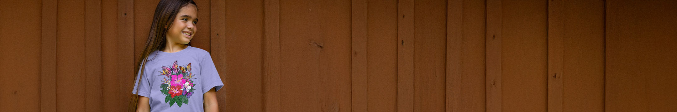 Young girl wearing a lavender shirt with a floral design on the front standing in front of a wood wall
