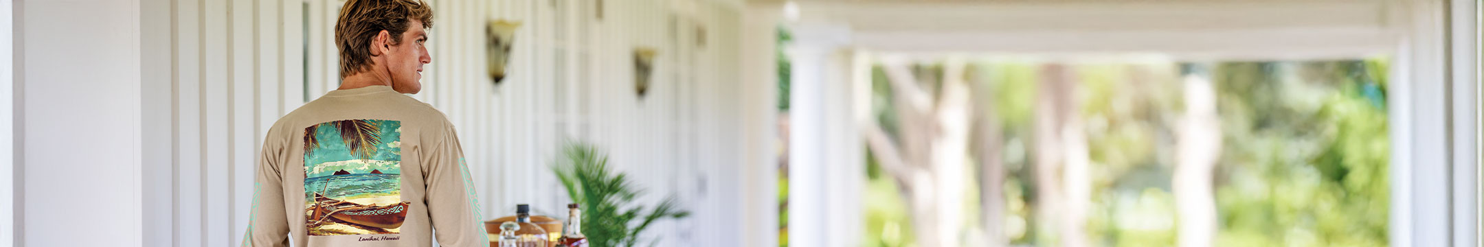 Man wearing a tan long sleeve with an image of Lanikai printed on the back, standing on the front patio of a house