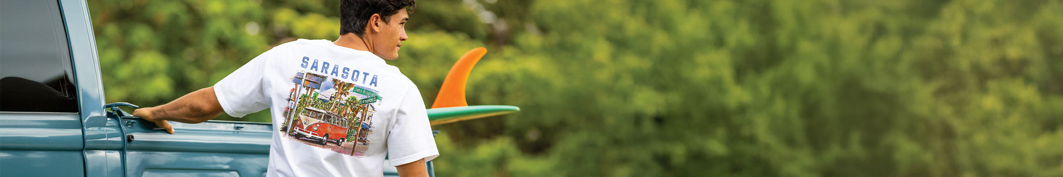 Person wearing a 'Sarasota' t-shirt with a surfboard, standing next to a vehicle in a natural setting.