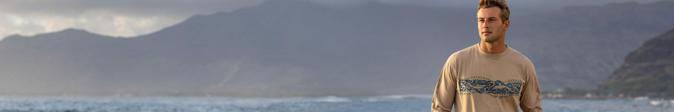 Man standing on a beach with mountains in the background wearing a kona coffee long sleeve
