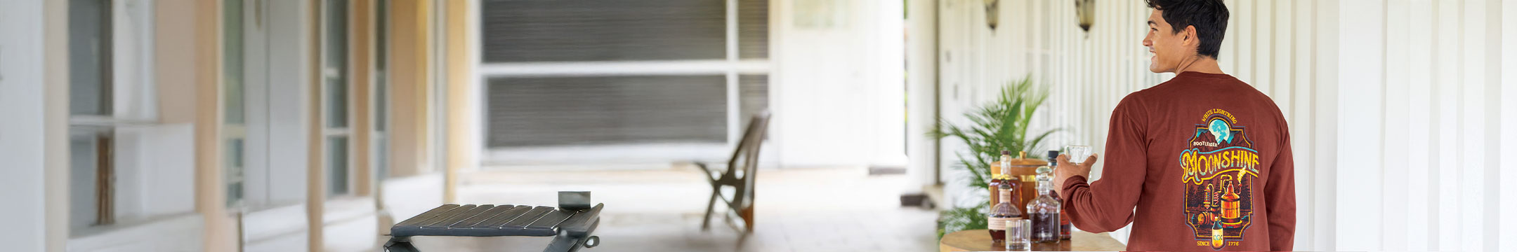 Person standing on a porch with a coffee table and chairs, holding a cup.