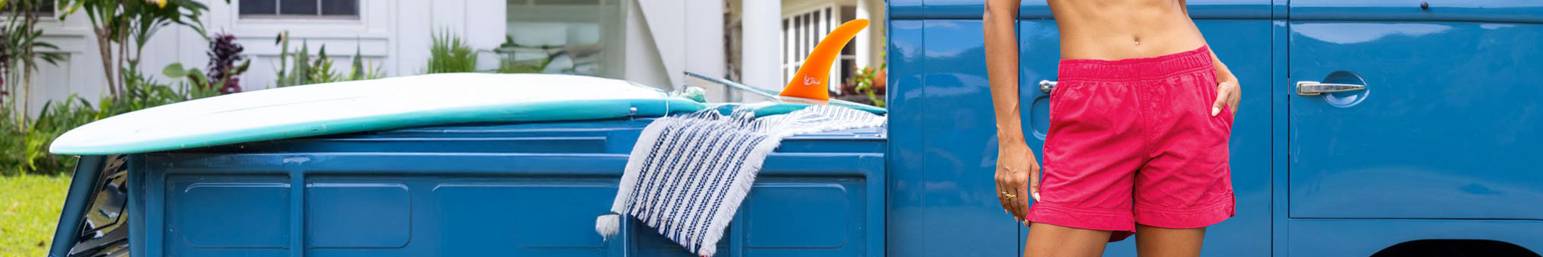 Person wearing Raspberry Dyed shorts standing next to a blue vehicle with a surfboard on top.