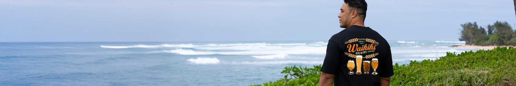 Man in a black t-shirt with a beer brand logo standing on a grassy area overlooking the ocean.