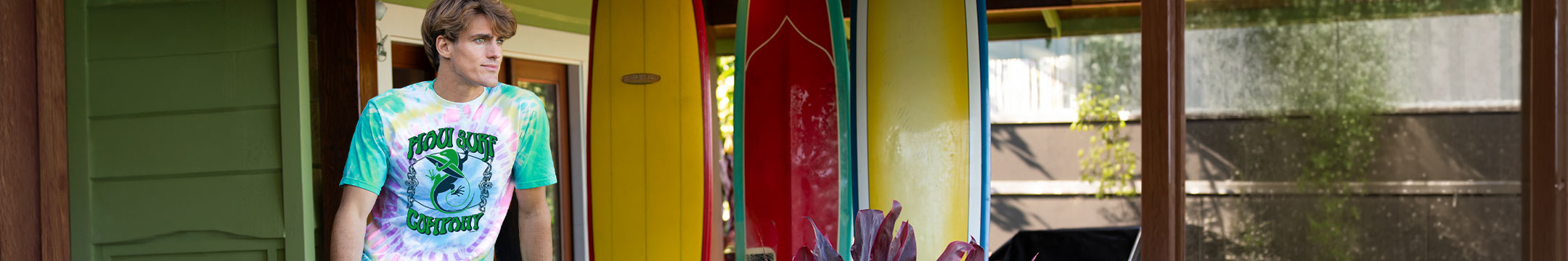 Man wearing a tie-dyed gecko t-shirt standing on a porch with surfboards in the background