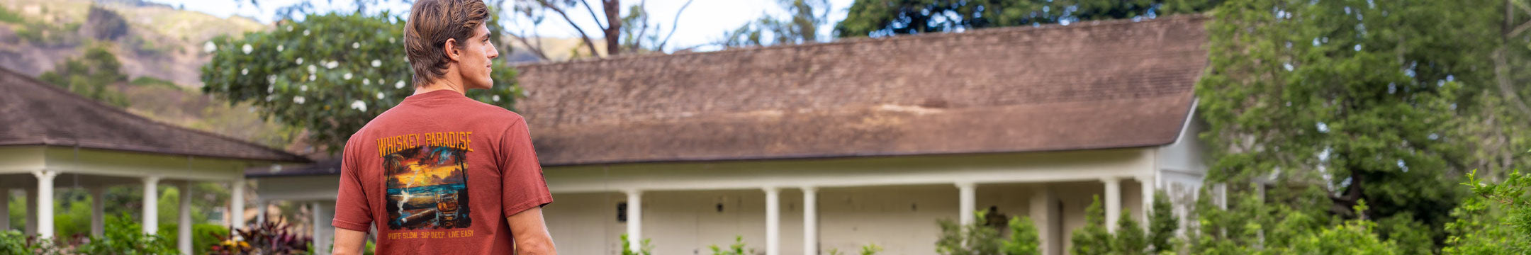 Man wearing a red shirt with a graphic design, standing in front of a house with a brown roof.
