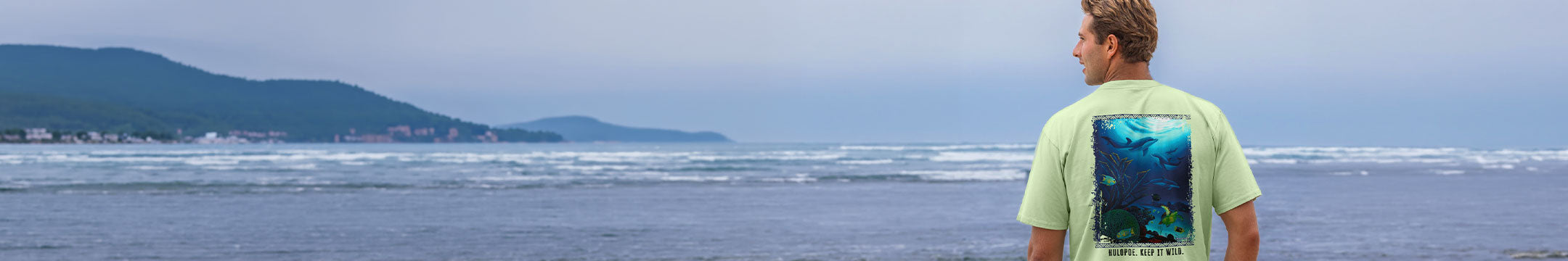 Person wearing a green t-shirt with a graphic design, standing on a beach looking at the ocean.