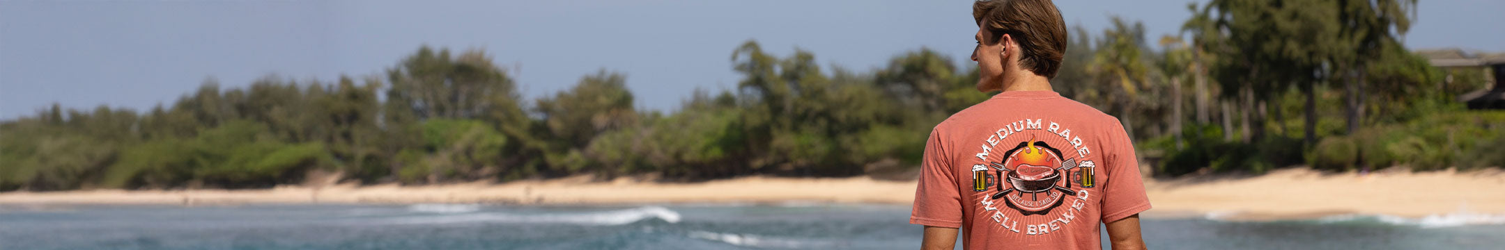 Person wearing a red t-shirt with a graphic design, standing on a beach with greenery in the background