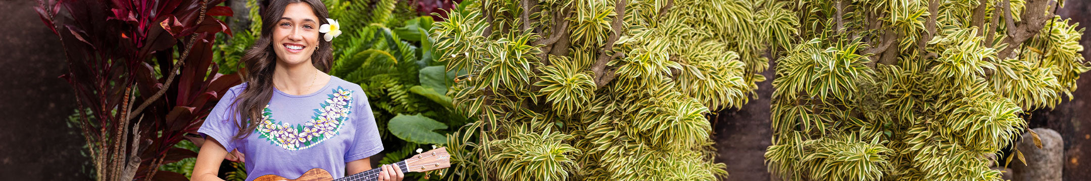 Woman wearing a purple t-shirt with a floral design playing an ukulele in front of green foliage.