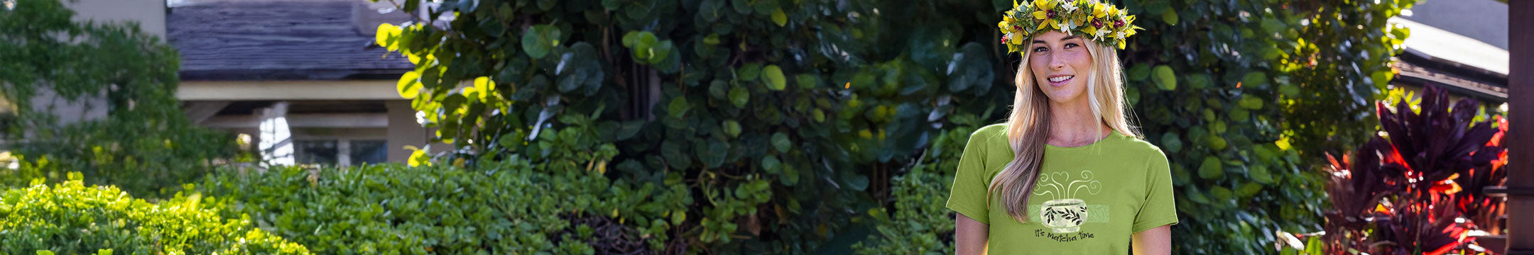 Woman wearing a green t-shirt with a logo, standing outdoors with greenery around.