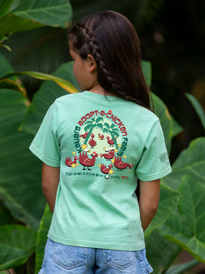 Child wearing a light green t-shirt with a graphic design, standing in front of large green leaves.