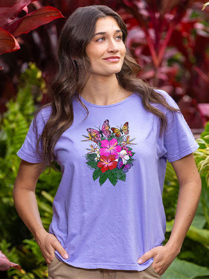 Woman wearing a purple t-shirt with a floral and butterfly design, standing in front of tropical plants.