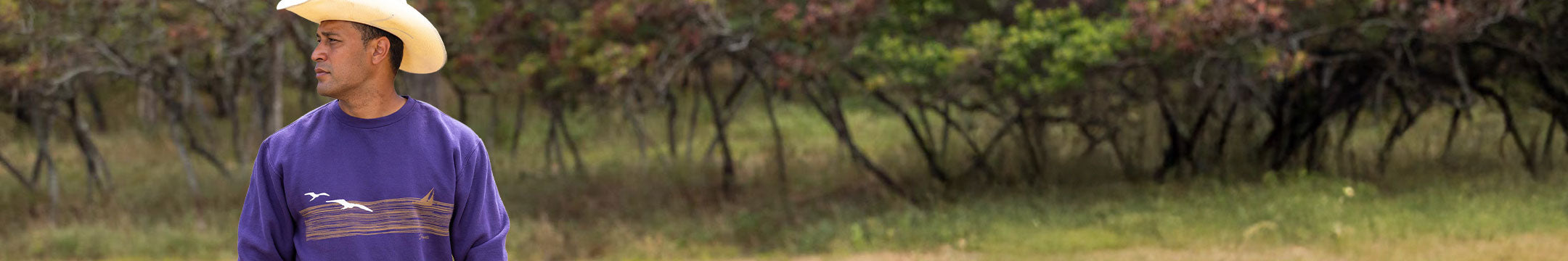 Person wearing a blackberry dyed sweatshirt and cowboy hat standing in a grassy field with trees in the background