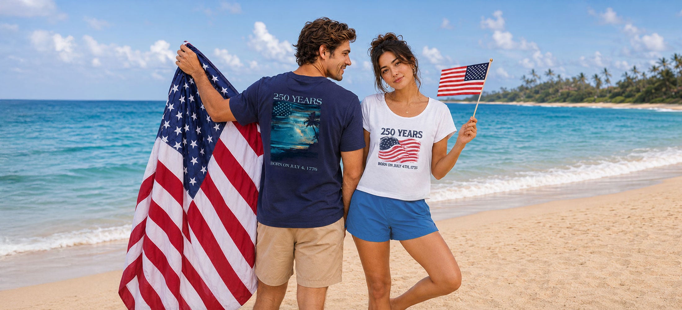 A couple standing on a sandy beach with the ocean behind them, each holding an American flag. The man wears a navy blue graphic T-shirt and khaki shorts, and the woman wears a white graphic T-shirt and blue shorts.