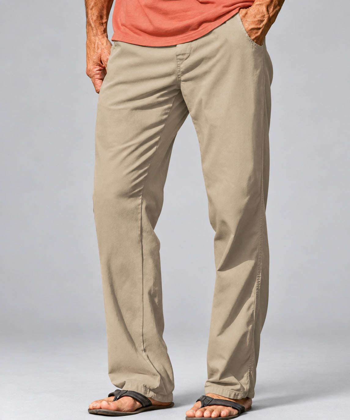 A person wearing straight-leg khaki pants, an orange t-shirt, and dark brown sandals, shown from the waist down against a light gray studio background.