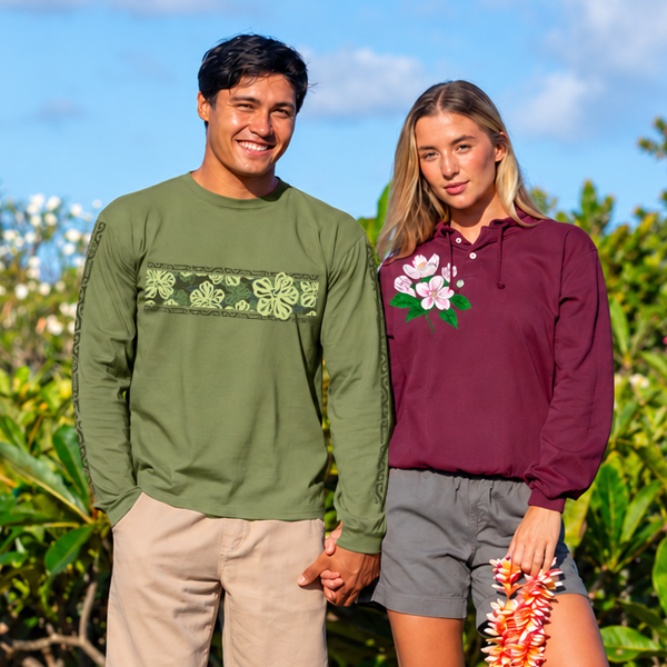 A man and a woman standing outdoors in a tropical garden setting, holding hands. The man wears an olive green long-sleeve shirt with a horizontal green floral pattern across the chest and beige pants.