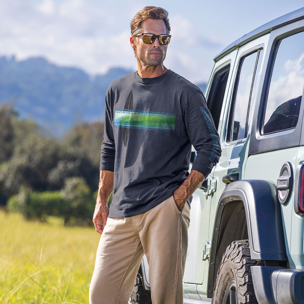 A man wearing a navy blue long-sleeve crewneck shirt with a colorful tropical palm tree graphic across the chest, paired with beige pants, standing outdoors next to a light blue vehicle with mountains in the background.