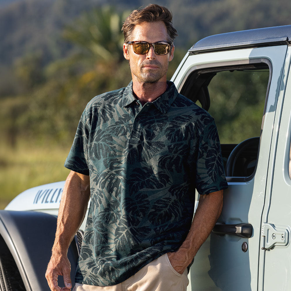 A man wearing a dark green tropical leaf print short-sleeve polo shirt and sunglasses, standing next to a light blue vehicle outdoors.