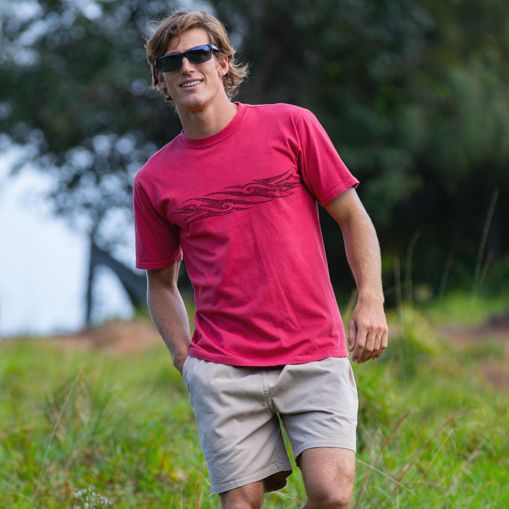 A man wearing a bright pink t-shirt with a dark graphic print and beige shorts, walking outdoors in a grassy field with trees in the background.