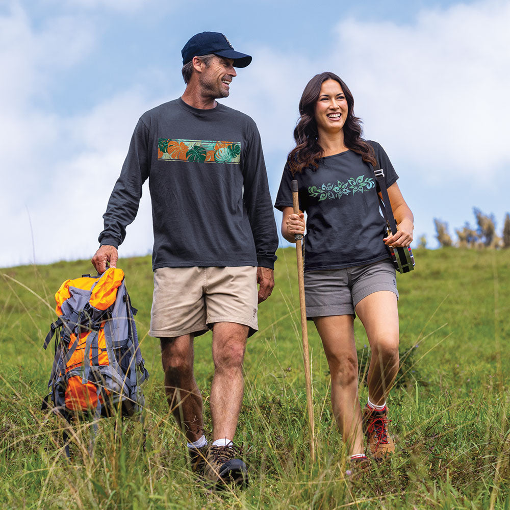 A man and a woman hiking outdoors on a grassy hill under a blue sky. The man wears a navy long-sleeve shirt with a colorful chest graphic, khaki shorts, and carries a large hiking backpack.
