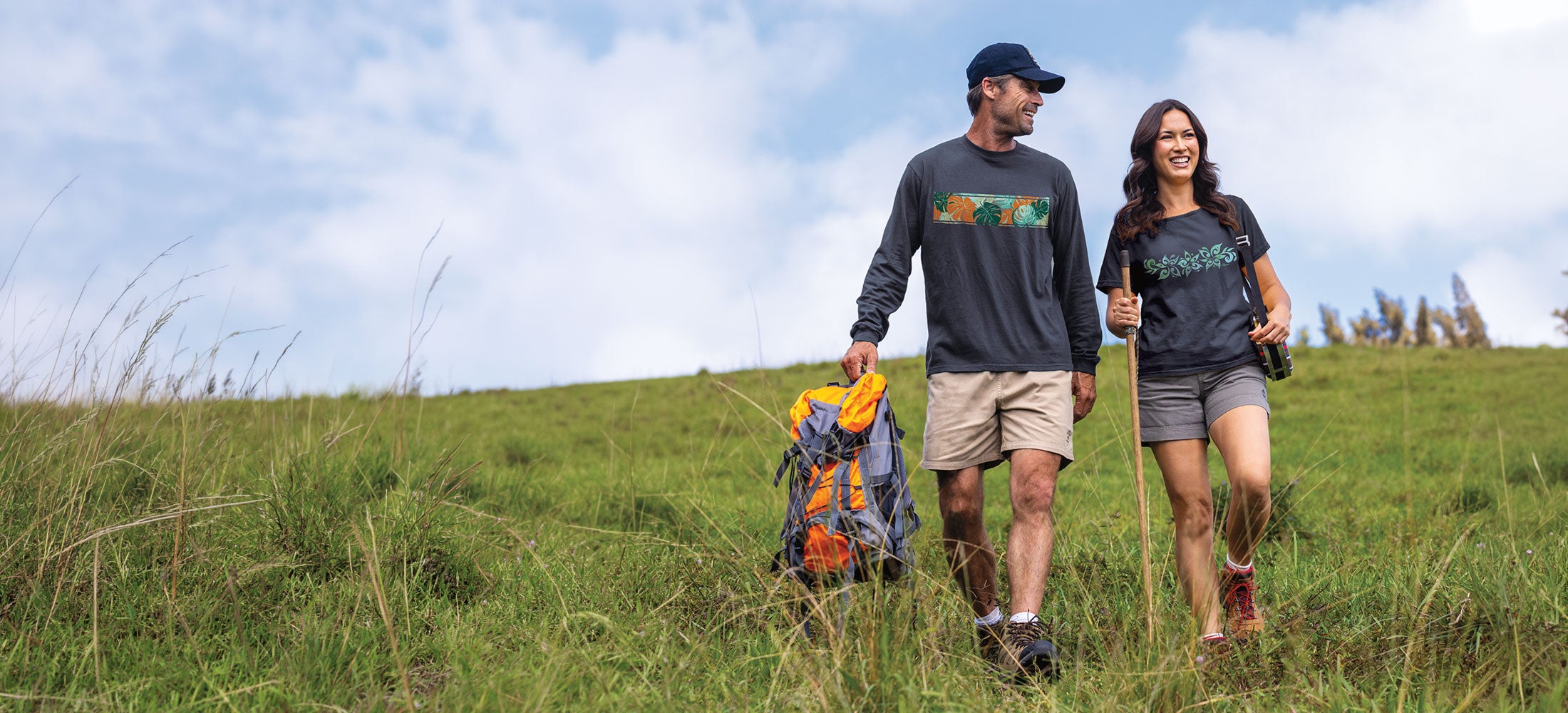 A man and a woman hiking outdoors on a grassy hill under a cloudy sky, both wearing matching navy blue long-sleeve shirts with a colorful graphic across the chest.