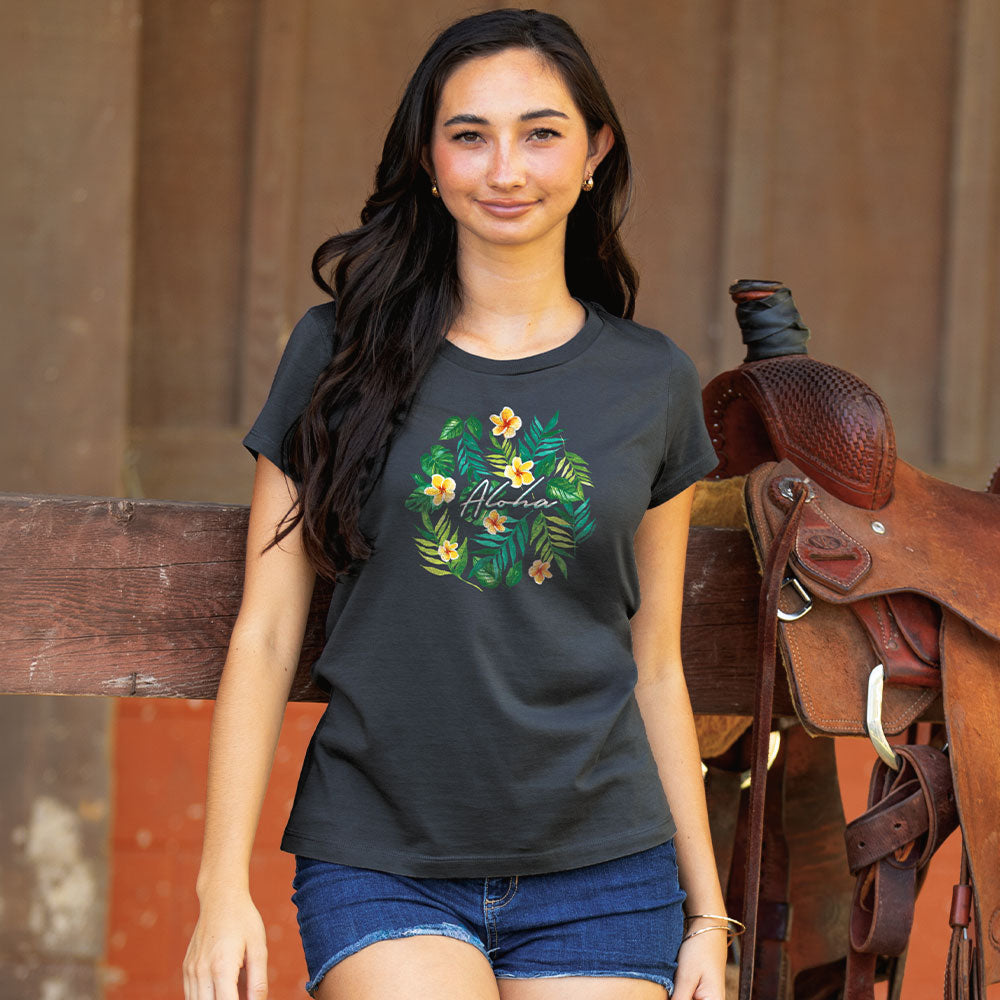 A woman wearing a dark charcoal gray t-shirt with a green tropical floral wreath graphic and the word 'Mistral' in script, paired with denim shorts, standing next to a brown leather western saddle.