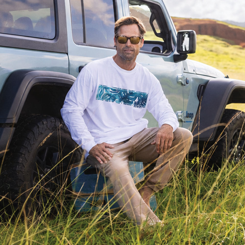 A man wearing a white long-sleeve crewneck shirt with a teal and blue tropical palm leaf graphic across the chest, khaki pants, and sunglasses, sitting on a blue cooler in front of a light blue Jeep in a grassy outdoor setting.