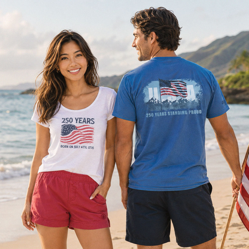 A man and a woman standing on a sandy beach holding hands, wearing matching patriotic t-shirts that celebrate 250 years of the United States. The woman wears a white t-shirt with a red and blue American flag graphic, and the man wears a blue t-shirt with a white and red American flag graphic on the back.