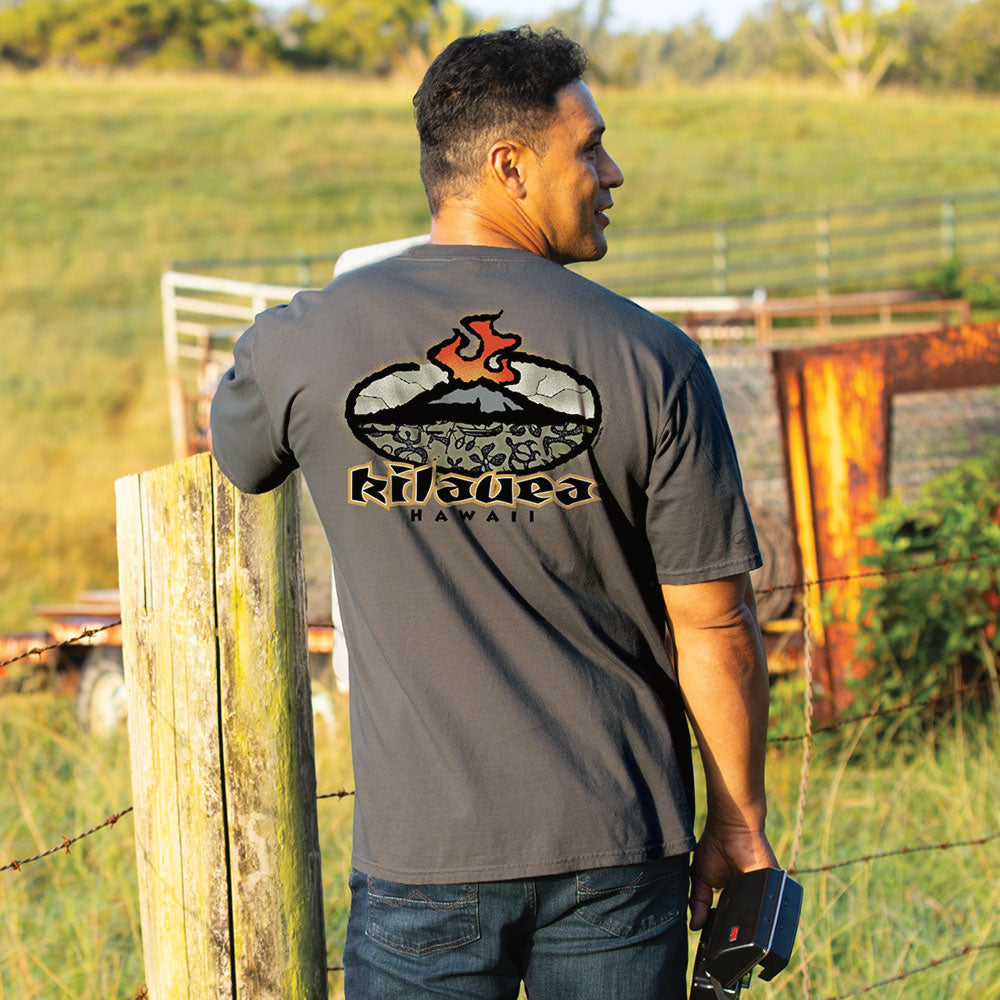 A man wearing a dark gray t-shirt with a 'Kilauea Hawaii' volcanic graphic on the back, standing outdoors in a grassy field near a wooden fence.