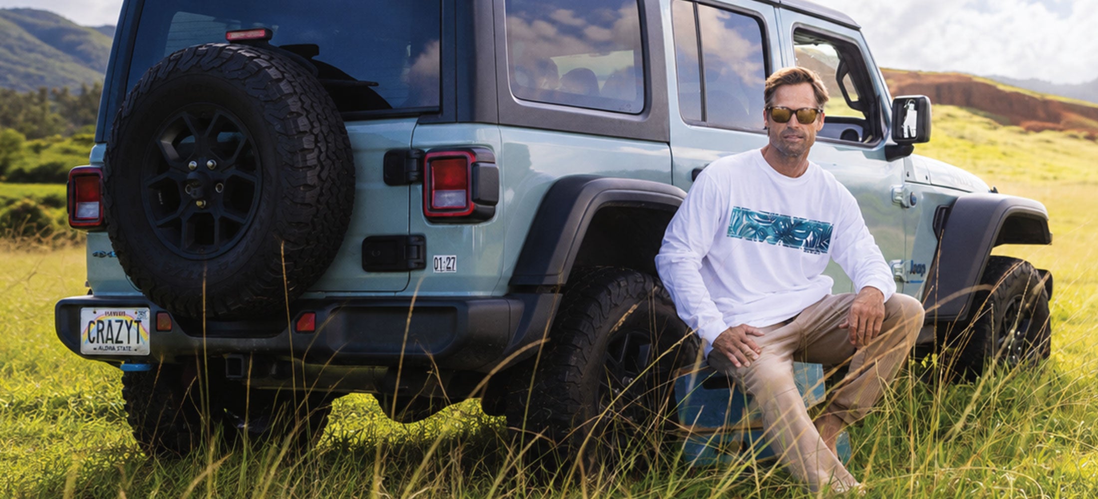 A man wearing a white long-sleeve t-shirt with a blue graphic and khaki pants sits in a grassy field next to a light blue Jeep Wrangler.