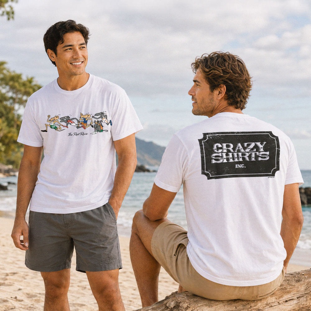 Two men on a beach wearing white graphic t-shirts. The standing man wears a white t-shirt with a colorful rowboat graphic and the seated man wears a white t-shirt with a large black 'CRAZY SHIRTS ENG.