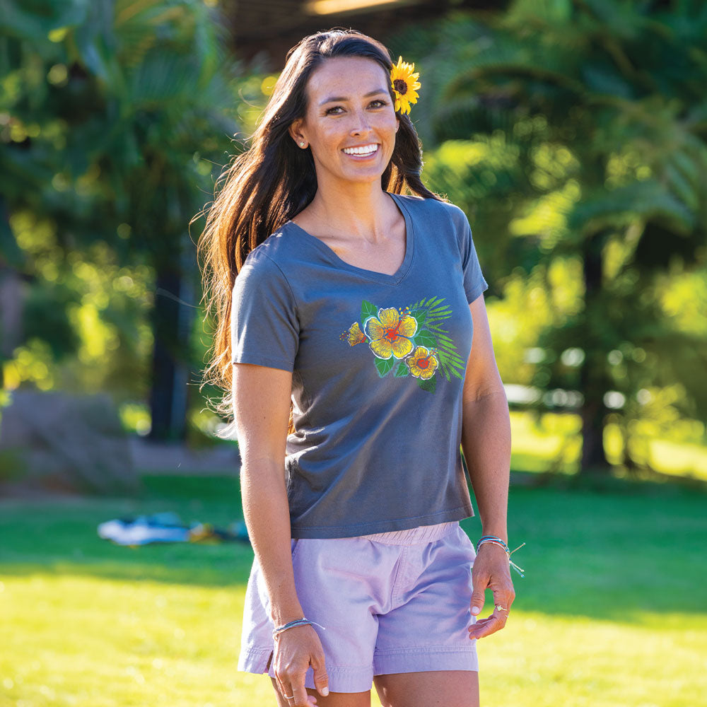 A woman standing outdoors in a sunny park wearing a dark gray v-neck t-shirt with a colorful hibiscus flower graphic on the front, paired with light lavender shorts.