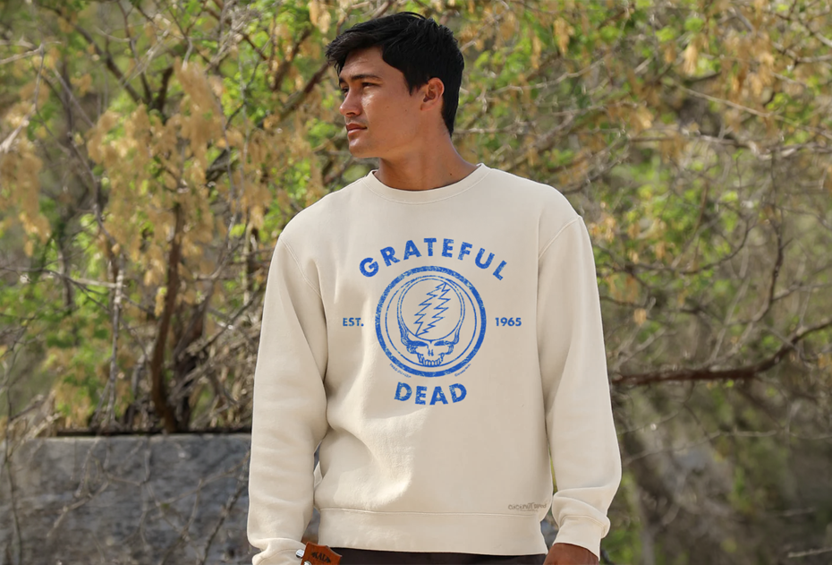 A man wearing a cream-colored Grateful Dead sweatshirt with a blue graphic print, standing outdoors in front of a blurred background of trees and branches.