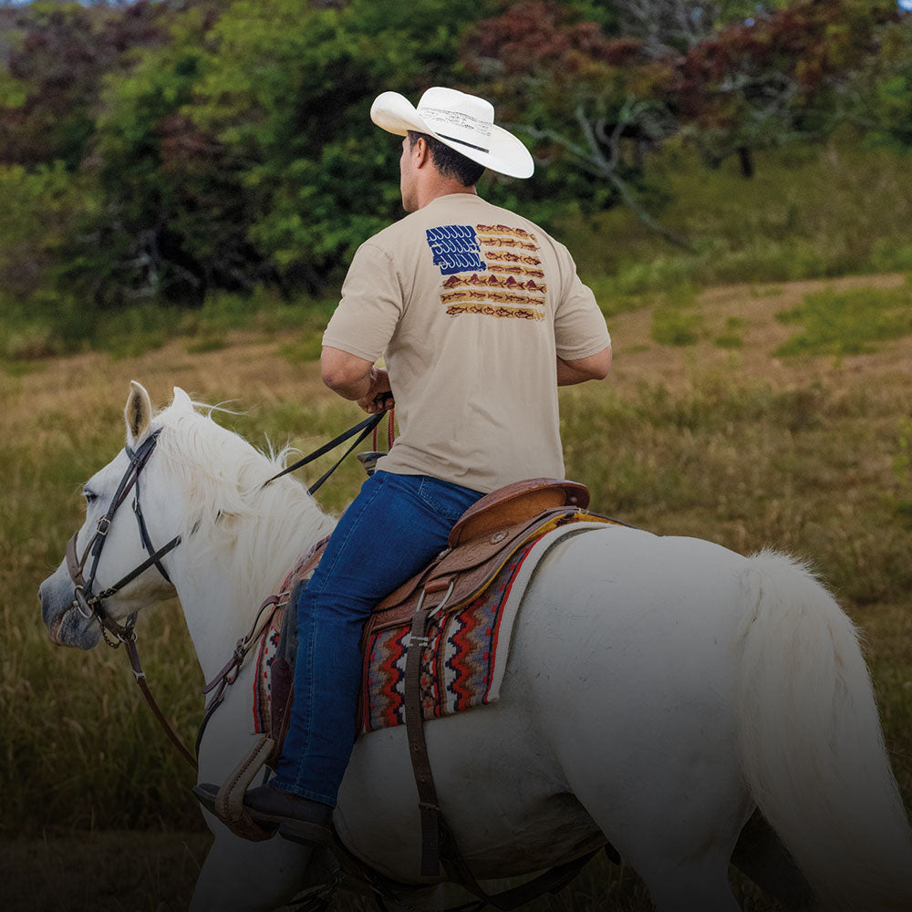 Man riding a white horse in a grassy field wearing a beige shirt with a logo.