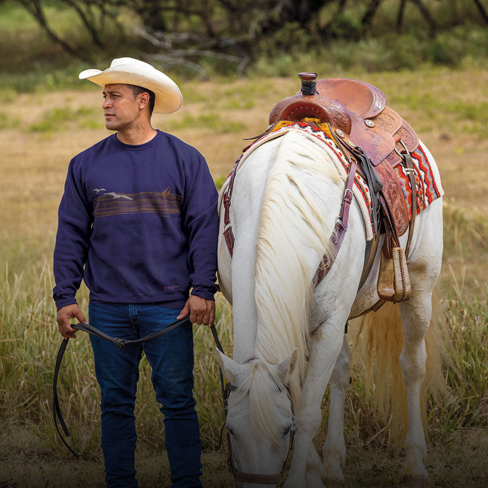 Man in a cowboy hat standing next to a white horse with a saddle in a field