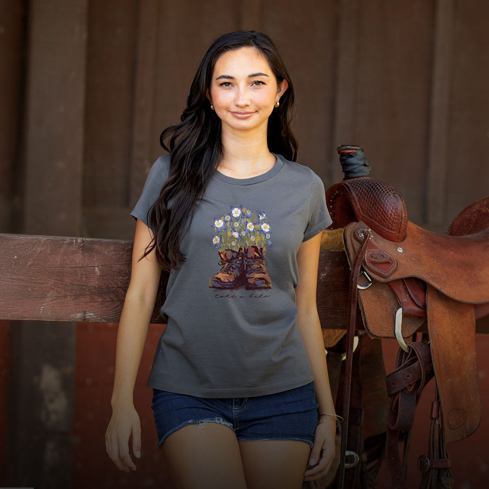 Woman wearing a t-shirt with a graphic design of boots and flowers, standing next to a horse saddle.