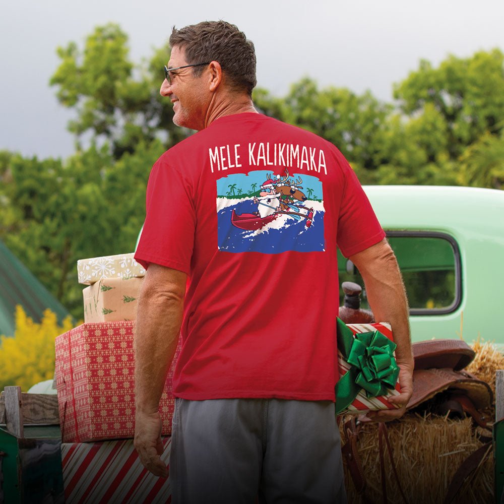 Man wearing a red shirt with a graphic design, standing outdoors with greenery and a vintage truck in the background.
