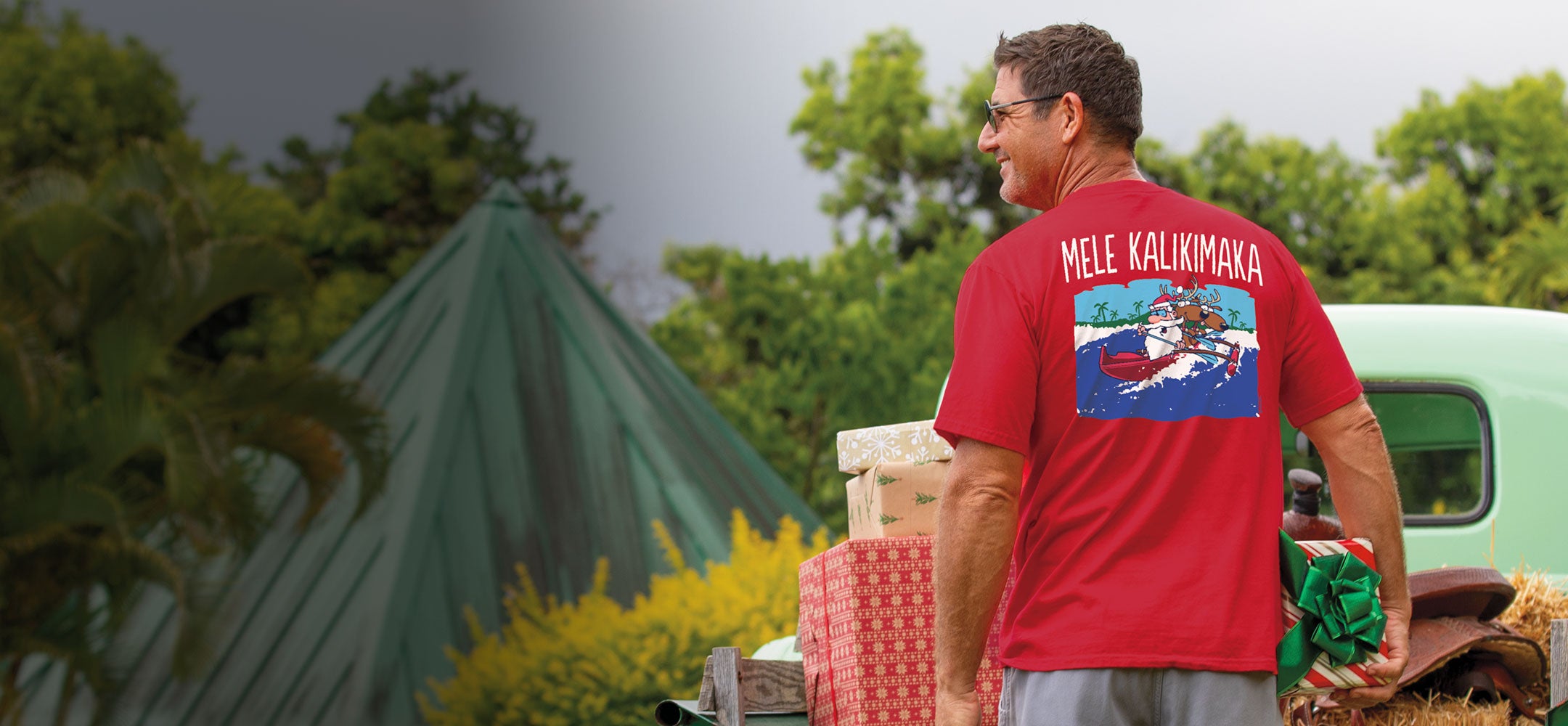 Man wearing a red t-shirt with a graphic design, standing outdoors with greenery and a vintage car in the background.