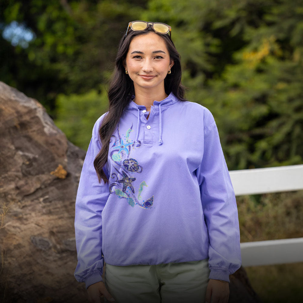 Woman wearing a purple hoodie with a nature-themed design, standing outdoors with greenery in the background.