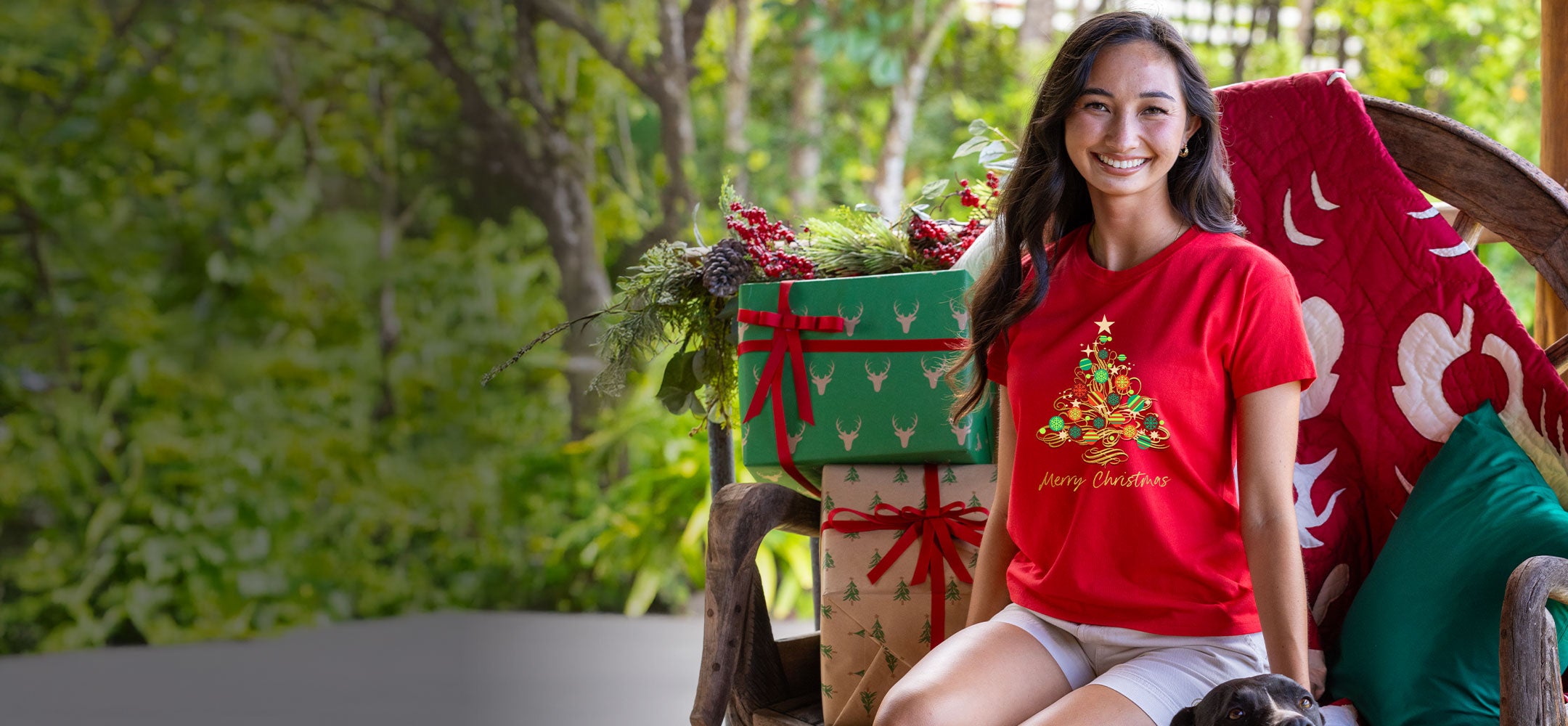 Woman wearing a red Christmas-themed t-shirt sitting on a wooden chair with decorative items in the background.
