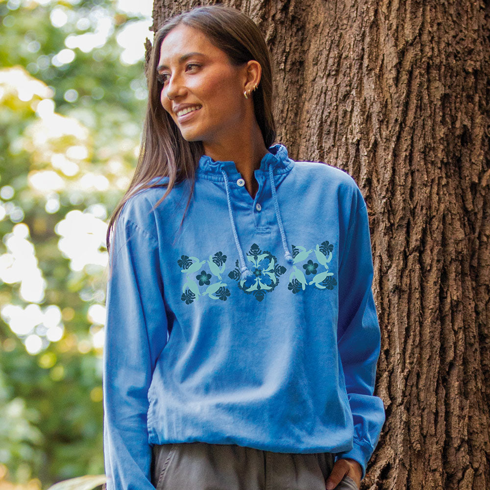 A woman wearing a light blue long-sleeve hooded top with a floral pattern across the chest, standing outdoors next to a large tree trunk.