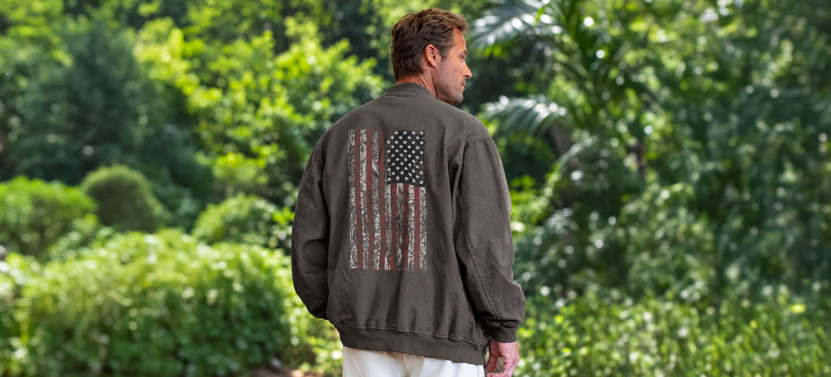 A man viewed from behind wearing a dark brown bomber jacket with a distressed American flag graphic on the back, standing in a lush green outdoor setting.