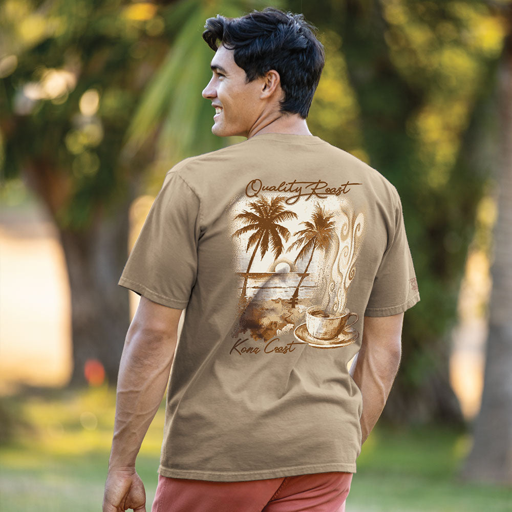 A man wearing a tan graphic t-shirt with a Hawaii-themed design on the back, standing outdoors in a park with blurred trees in the background.