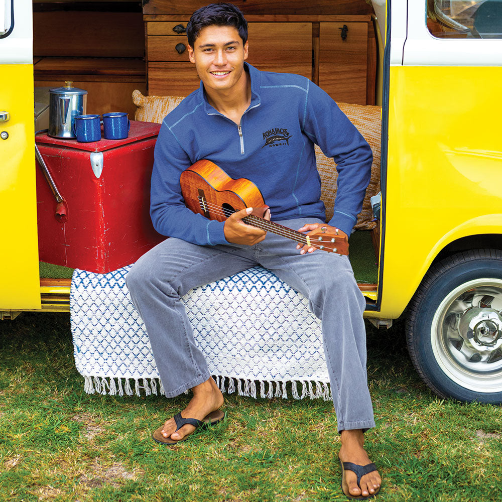 A man sitting on a white and blue patterned blanket in front of a yellow vintage camper van, wearing a navy blue quarter-zip pullover, gray pan, and black sandals, while holding a wooden ukulele.s