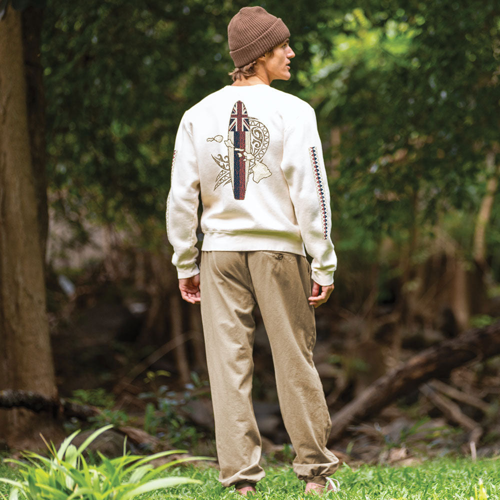 A man standing outdoors in a wooded area, viewed from behind, wearing a cream sweatshirt with a large surfboard graphic on the back, olive pants, and a brown beanie.