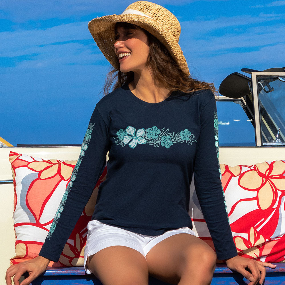 A woman wearing a navy long-sleeve top with a teal floral chest print and matching floral sleeve cuff, seated outdoors on a boat with colorful pillows and blue sky in the background.