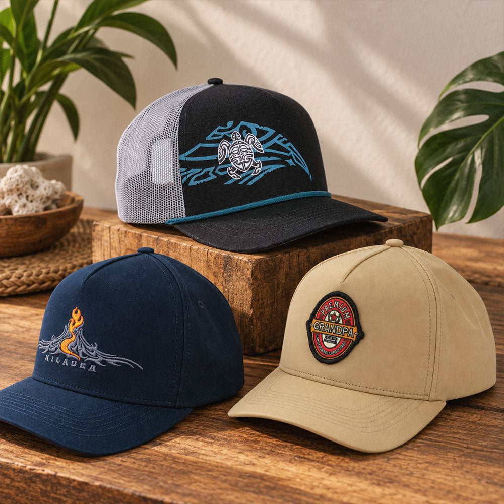 Three embroidered trucker-style caps displayed on a wooden surface with plants in the background. The top cap is black and gray with a blue turtle graphic; the bottom-left cap is navy; the bottom-right cap is tan with a circular patch.