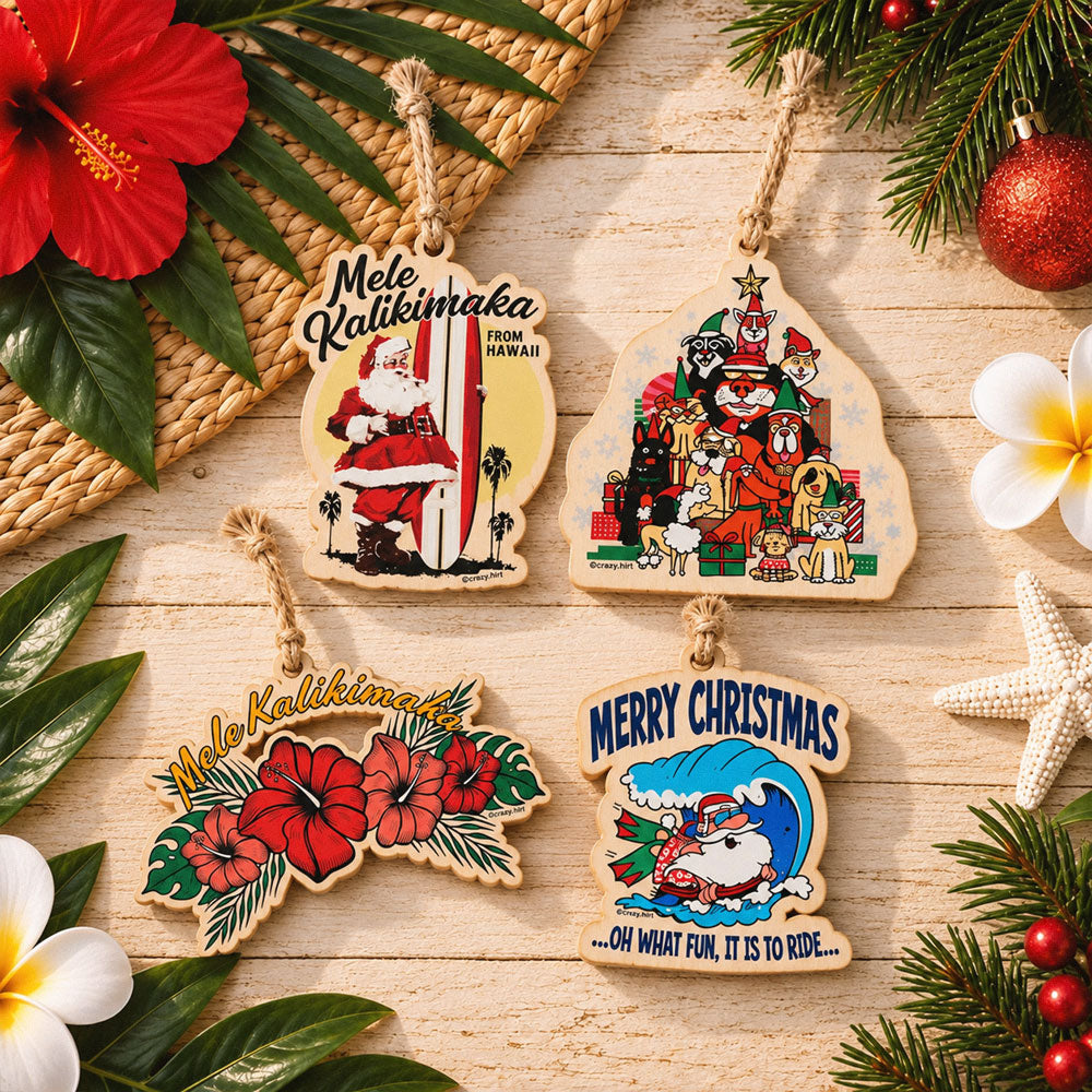 Five wooden Christmas ornaments with Hawaiian-themed illustrations and text, arranged on a light wood surface with tropical flowers, greenery, a woven mat, and holiday decorations around them.