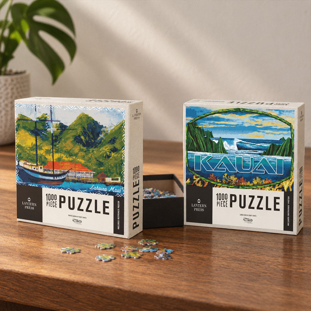 Two Lantern Puzzle 1000 Piece Puzzles on a wooden table. The left puzzle box features an illustration of a sailboat on a tropical lake, and the right puzzle box features an illustration of the Kauai landscape.