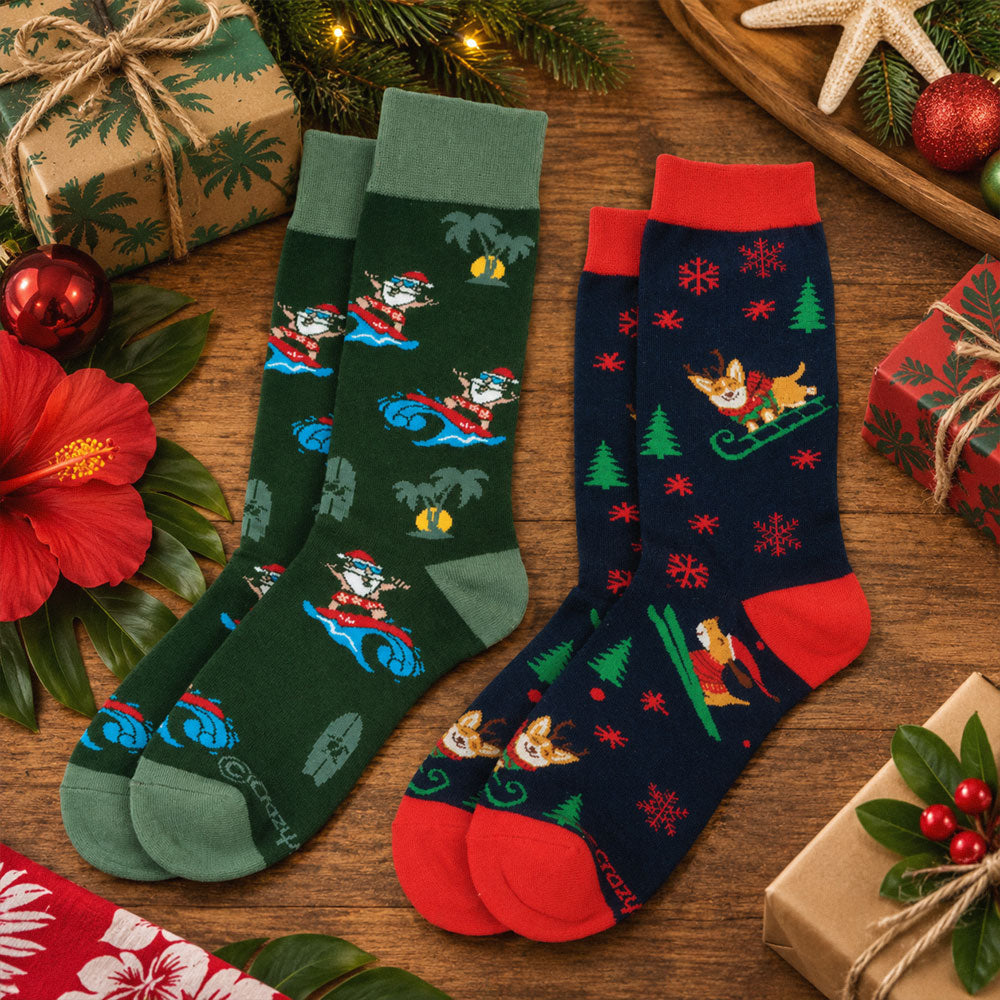 Two pairs of Christmas-themed crew socks displayed on a wooden surface with holiday decorations. One pair is dark green with Santa and snowman motifs, and the other is dark navy with Santa and dog motifs.
